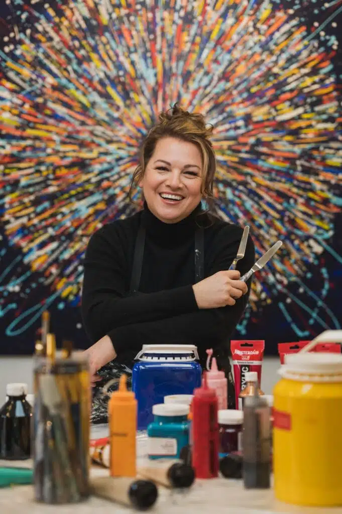 Quebec artist Caroline Bergeron (ARO) smiling in her studio, seated in front of a large, brightly colored abstract acrylic painting with art supplies and paints nearby.
